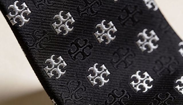 Black silk tie with silver crest embroidery, diagonal closeup showing twill ribbed texture