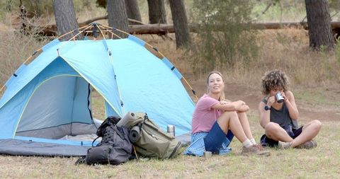 Two Friends Relax Near Tent in Scenic Forest Setting, Enjoying Nature Camping
