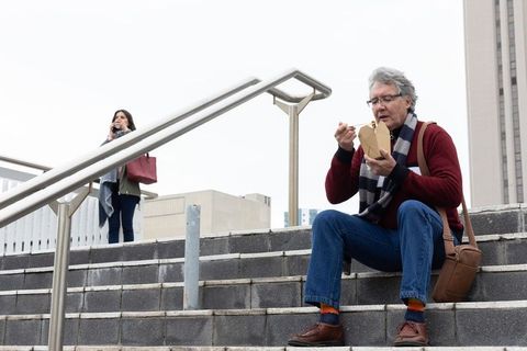Senior man eating takeout on urban steps while woman talking on smartphone in plaza overcast