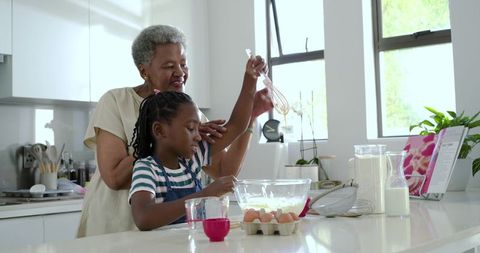 Grandmother guiding grandchild baking in bright kitchen whisking batter together