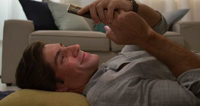 Image showing a man lying on the floor using a smartphone at home, with a relaxed and smiling expression. Perfect for depicting concepts like home comfort, leisure time, relaxation, and mobile technology usage. Suitable for advertisements, lifestyle blogs, and social media content related to modern living, technology, and relaxation.