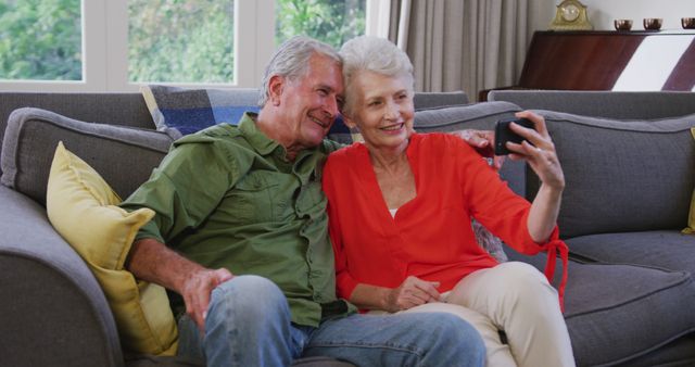 Older couple sitting on a grey sofa in a living room, smiling and taking selfies with a smartphone. Could be used for topics related to seniors, modern technology, family bonding, retirement lifestyle, and indoor leisure activities.