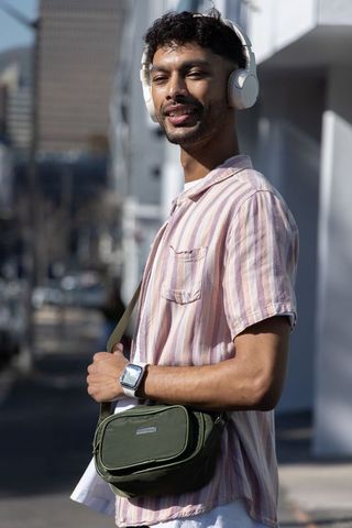 Indian man listening to music on headphones wearing pastel shirt and crossbody bag in sunny city
