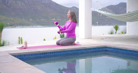 Senior woman practicing gentle dumbbell workout on terrace by pool with ocean view