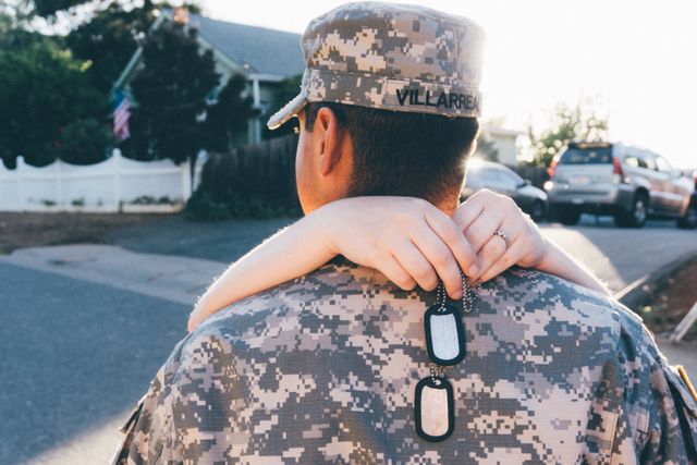 A soldier in a camo uniform stands outside a house while being embraced from behind by a loved one. Dog tags are visible, adding emotional depth to the scene. This image is perfect for illustrating themes of military life, emotional reunions, sacrifices, and the strong bonds between military personnel and their families, suitable for use in advertisements, articles about veterans, and family-oriented military support campaigns.