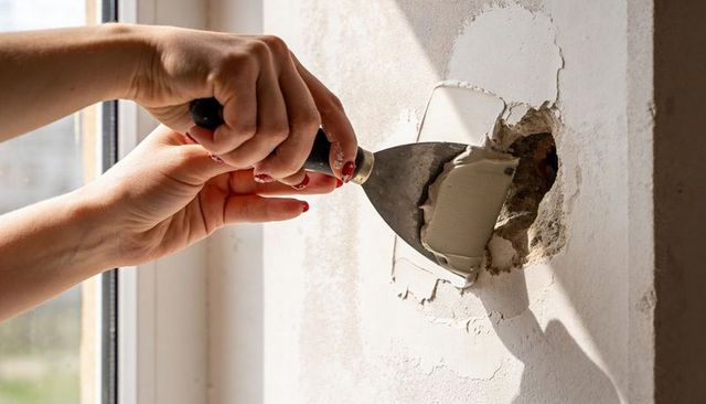 Hands repairing wall hole with putty knife applying plaster patch by window in light