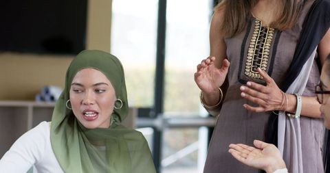Female colleagues collaborating in modern office, woman wearing green hijab sharing ideas