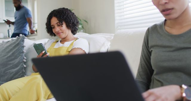 Three people in a cozy living room, using digital devices for various activities. Professional and personal uses of technology shown. Perfect for themes of modern work-life balance, home office setups, and collaboration.