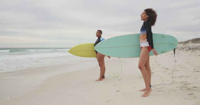 Happy african american female friends on the beach holding surfboards looking toward sea. healthy outdoor leisure time by the sea.