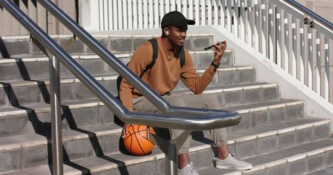 Young African American man sitting on urban steps holding basketball and using smartphone