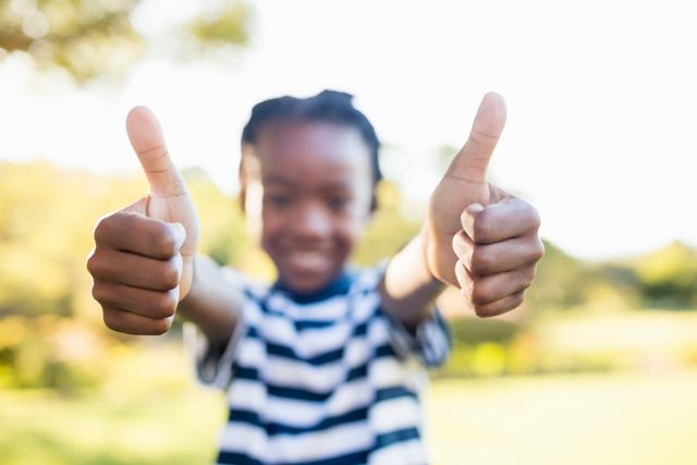 Boy giving thumbs up in park, smiling and enjoying sunny day. Perfect for themes of childhood joy, outdoor activities, positive emotions, and carefree moments. Suitable for use in advertisements, educational materials, and family-oriented content.