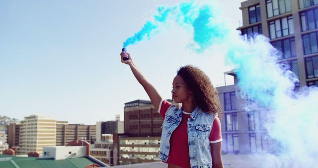 Young biracial woman holds a blue smoke flare outdoors, with copy space. She exudes confidence against an urban backdrop.