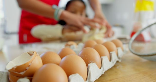 A mother and child are seen baking together in a kitchen, with the focus on a carton of eggs in the foreground. This image can be used in articles or advertisements related to family activities, cooking tutorials, parent-child bonding, or culinary blogs. It evokes feelings of warmth, togetherness, and home cooking.