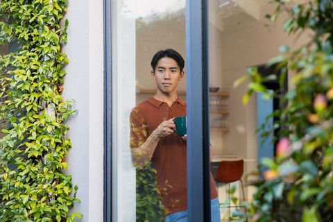 Asian man sipping coffee by sliding glass door with climbing vines and modern kitchen