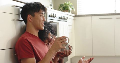 Mixed-race couple sitting on kitchen floor holding mugs, sharing cozy sunlit morning