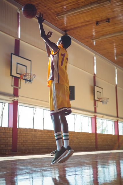 Teenage boy in yellow basketball uniform jumping to shoot a basketball in an indoor court. Ideal for use in sports-related content, youth athletic programs, fitness promotions, and team sports advertisements.