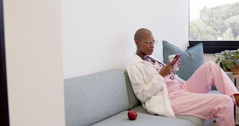 African clinician relaxing on clinic sofa in pink scrubs and lab coat using smartphone