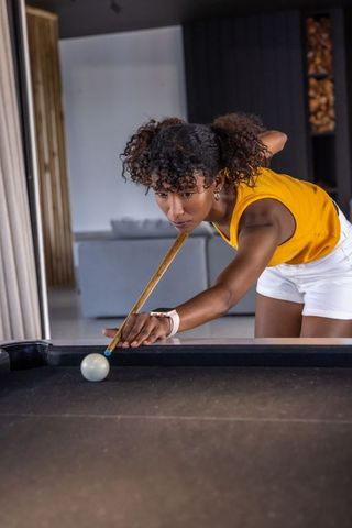 Young woman leaning and aiming cue on dark felt pool table in modern lounge interior