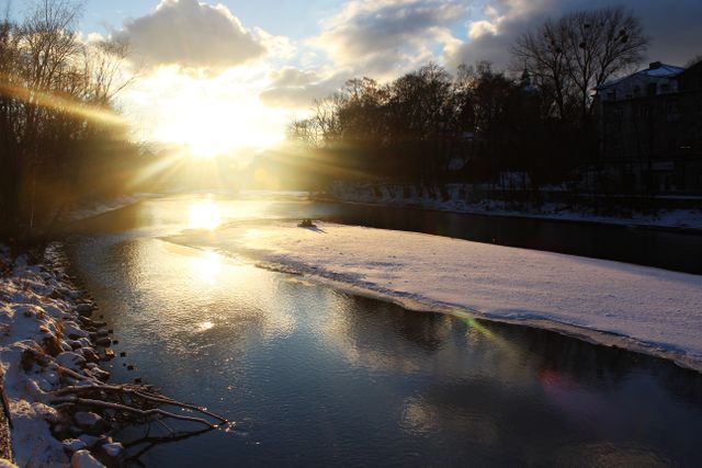 Sunset creates a beautiful glow over a tranquil winter river with icy patches and snow-covered banks. Trees line the river, casting shadows as the sun sets, making the scene serene and peaceful. Ideal for nature, winter, and tranquility-themed projects, suitable as background, screensaver, or for seasonal marketing materials.