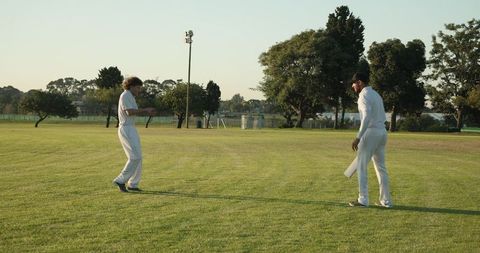 Cricketers Standing on Sunlit Field Wearing White Kit Holding Bat and Ball