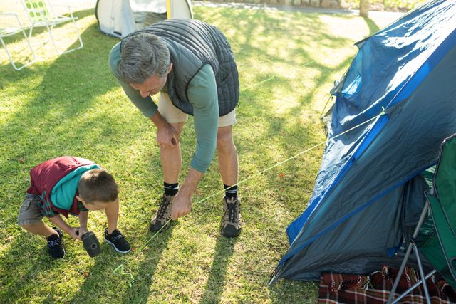 Grandfather showing grandson how to set up a camping tent on a grassy area. Ideal for use in articles or advertising about family activities, camping trips, outdoor education, or multigenerational bonding.