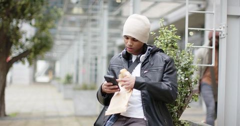 African american man checking smartphone while eating sandwich in urban plaza