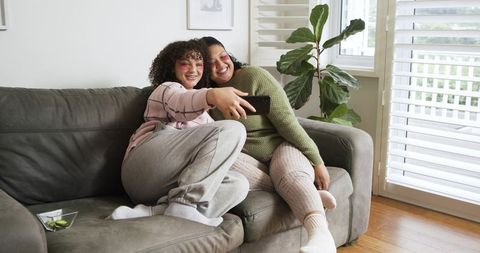 Mother and daughter taking selfie on gray sofa in cozy loungewear, smiling together