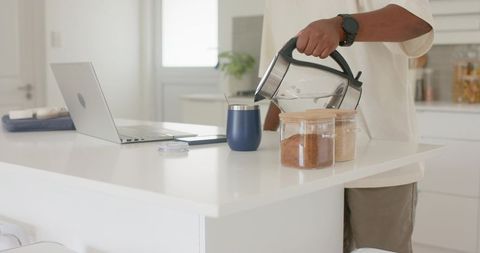 Pouring hot water into insulated tumbler beside laptop on modern white kitchen island