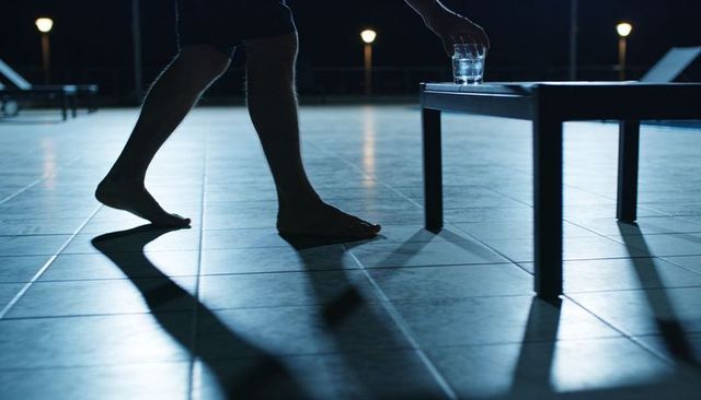 Barefoot man walking on tiled pool deck at night reaching for glass on low table