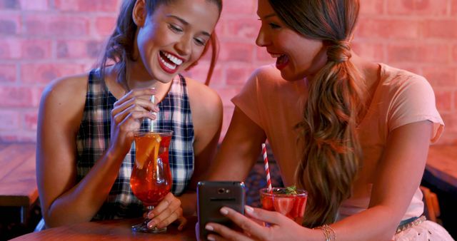 This image features two young women enjoying beverages at a trendy bar while looking at a smartphone. Both of them are engaged and laughing, suggesting they are possibly browsing through social media or looking at photos. The vivid background with brick walls and relaxed atmosphere makes it ideal for use in advertising for bars, nightlife promotions, social media campaigns, or articles about modern friendship and social interactions.