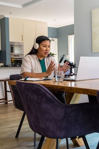 Middle-aged asian woman recording podcast at home wearing headphones speaking into mic