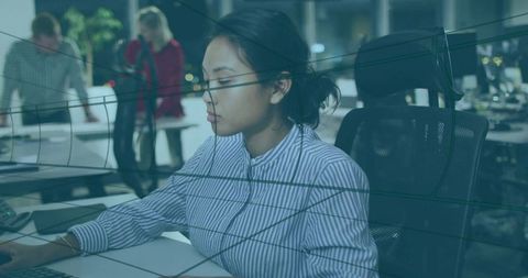 Focused woman typing in modern open-plan office with glass reflections and teamwork