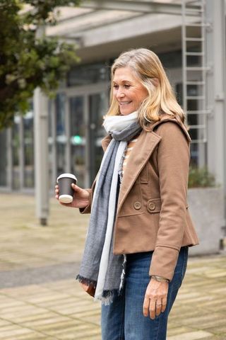 Senior woman standing and smiling while holding takeaway coffee near modern glass building