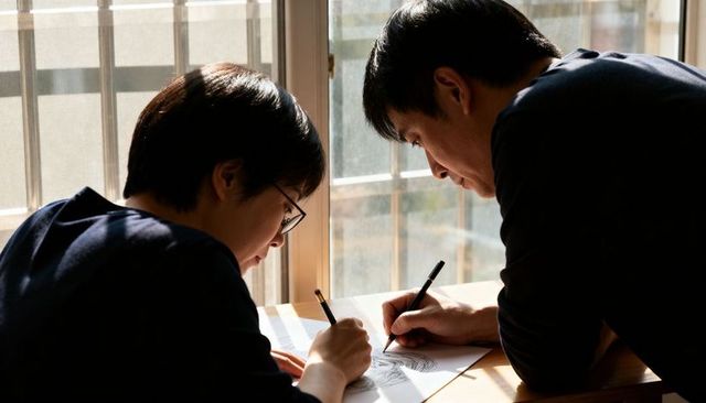 Mentor and student collaborating on pencil sketches at sunlit wooden table