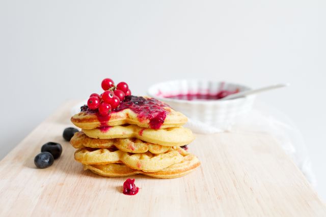 Stack of fluffy pancakes with vibrant berry sauce and garnished with red currants. Blueberries scattered on wooden board. Ideal for illustrating breakfast recipes, food blogs, and culinary websites.