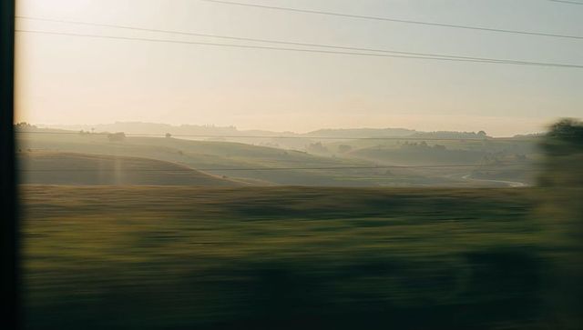Passing rolling fields and winding country road from moving vehicle at golden hour haze