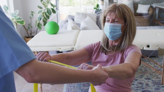 This scene shows a female health worker guiding a senior woman through exercise with a resistance band in a home setting, emphasizing at-home fitness, care, and precautionary measures with the use of masks. Ideal for use in contexts related to senior health care services, in-home physical rehabilitation, exercises for the elderly, or home wellness practices.