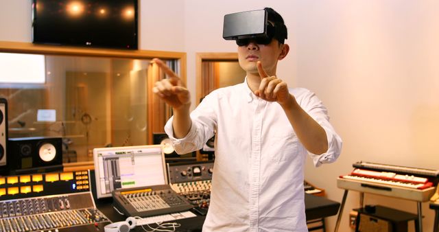 Man is using virtual reality headset while standing in music studio. Multiple audio equipment pieces and computers are visible in the background. Suitable for concepts related to modern music production, technology integration, virtual reality applications in creative industries, and innovative tools in music creation.