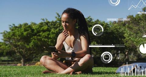 Young woman sitting cross-legged on grass checking smartphone with fitness hud overlays