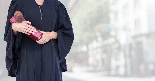 Judge in robe holding book and gavel, standing outdoors against blurred city street background. Ideal for content related to legal professions, judiciary, justice system, authority, government-related matters, and urban settings.