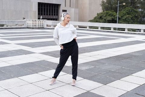 Mature woman stretching on urban checkerboard plaza wearing headphones and joggers