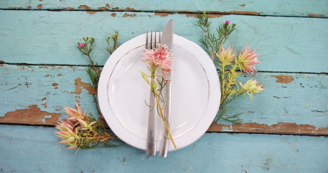 A white plate with silverware and delicate flowers is set on a rustic blue wooden table, with copy space. It evokes a sense of a spring or summer meal in a casual, charming setting.