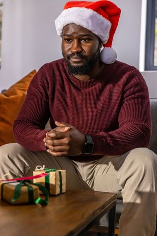 African american man wearing santa hat and maroon sweater sitting by gifts for holiday