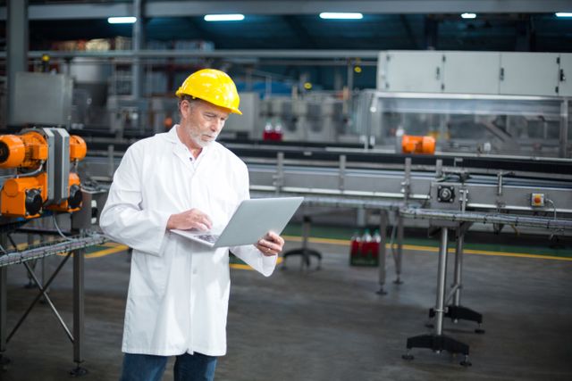 Factory engineer wearing safety helmet and lab coat using laptop in beverage production plant. Ideal for illustrating industrial operations, quality control processes, and modern manufacturing technology. Useful for articles on factory automation, engineering careers, and workplace safety.