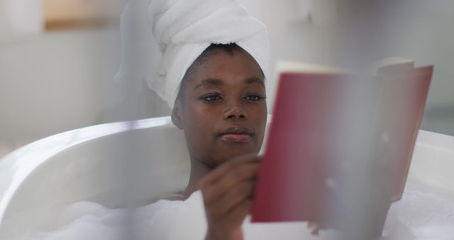 Portrait of african american attractive woman relaxing in bath and reading book in bathroom. beauty, pampering, home spa and wellbeing concept.
