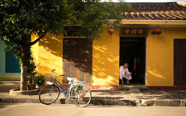 Depicts a tranquil street scene in an ancient town with a person sitting and reading outside a yellow building. Traditional elements like lanterns and a vintage bicycle enhance the calm atmosphere. Useful for illustrating concepts like cultural heritage, tranquil mornings, relaxation, traditional architecture, and leisurely activities in historical settings.