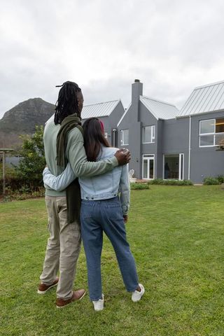Couple embracing on lawn in front of modern gray home with mountain backdrop and garden
