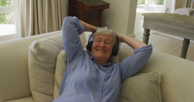 Senior woman with gray hair reclining on a sofa, wearing headphones and smiling. Ideal for projects related to relaxation, senior lifestyle, leisure activities, or promoting a relaxed and positive lifestyle for the elderly. Could also be used in healthcare contexts to highlight mental well-being or hear loss solutions.