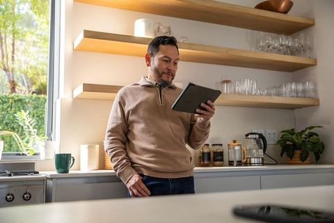 Mature asian man leaning in sunlit modern kitchen holding tablet and enjoying coffee
