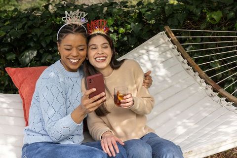 Friends celebrating outdoors on hammock wearing merry headbands and sharing phone with drink